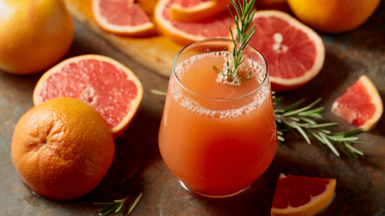 A glass of grapefruit juice with a rosemary sprig surrounded by whole and sliced grapefruit