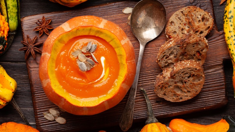 Bowl of pumpkin soup on a cutting board with bread surrounded by gourds and pumpkins