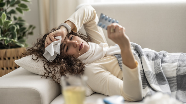 A sick woman lying on a couch holding a tissue and a blister pack of antibiotics