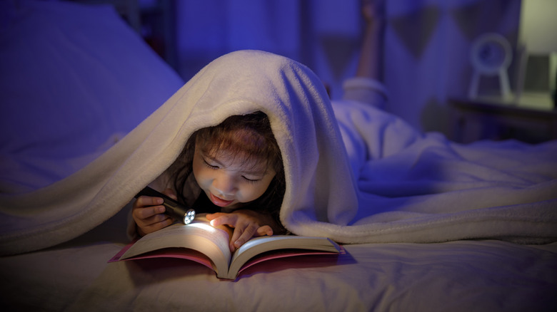 child under bed covers reading a book with a flashlight