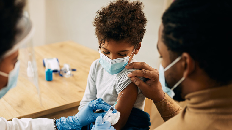 Young masked child receiving vaccine from masked doctor while sitting on his masked parent's lap