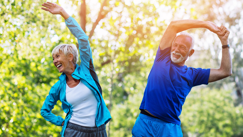 Older couple smiling and stretching outside in the sun wearing workout clothes