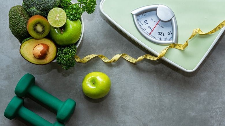 scale and measuring tape near dumb bells and bowl of green fruits