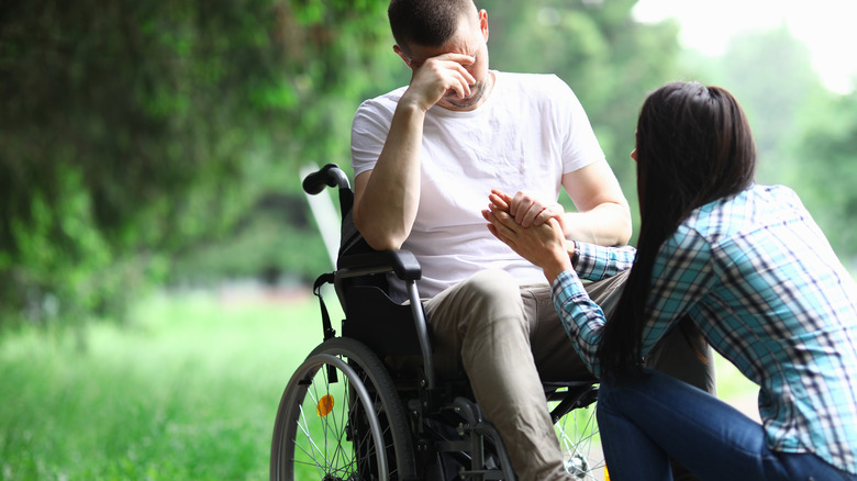 Distressed man in wheelchair while a woman kneels in front of him holding his hand