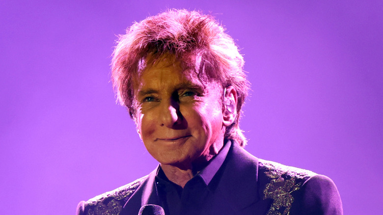 Barry Manilow seeped in a purple haze light smiling and holding a microphone.