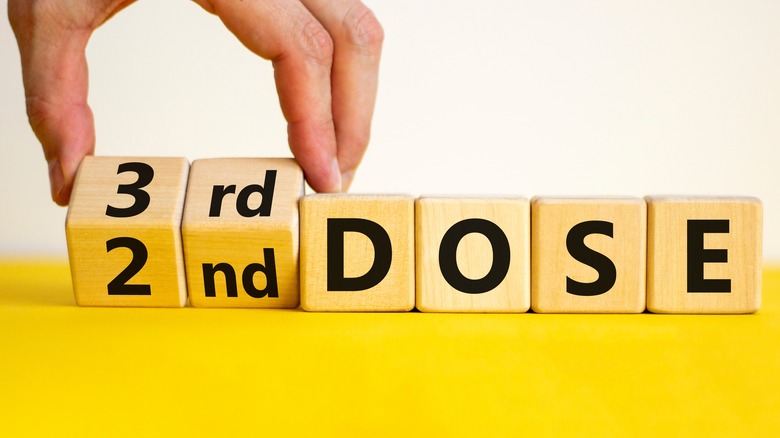 Hand flipping wooden block from reading "2nd DOSE" to reading "3rd DOSE" as it pertains to COVID vaccines