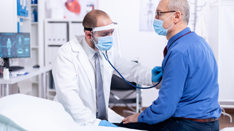 Masked doctor using stethoscope on masked senior patient