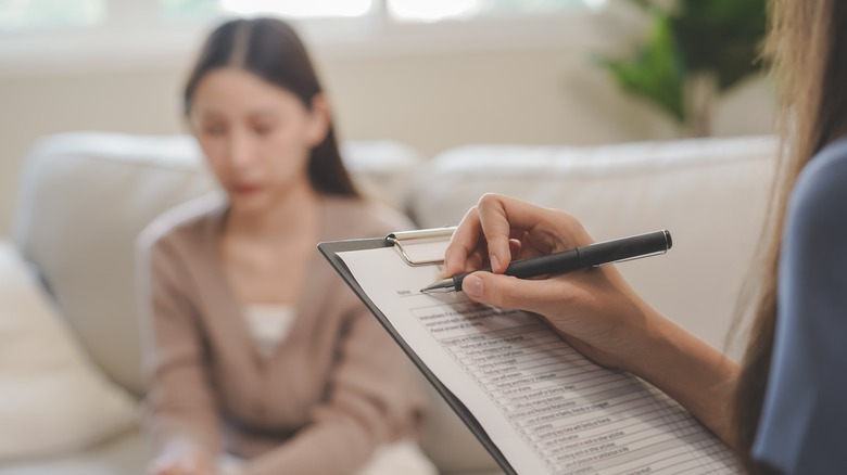 Therapist filling out paperwork on clipboard with blurred patient sitting on couch in background