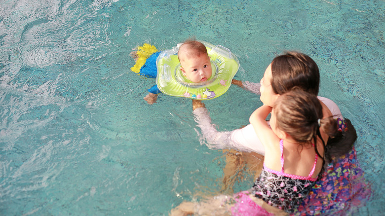 Baby in a pool wearing a neck float, with mom and sister