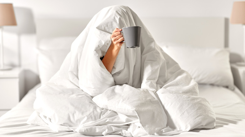 woman hiding in her comforter holding out a coffee cup