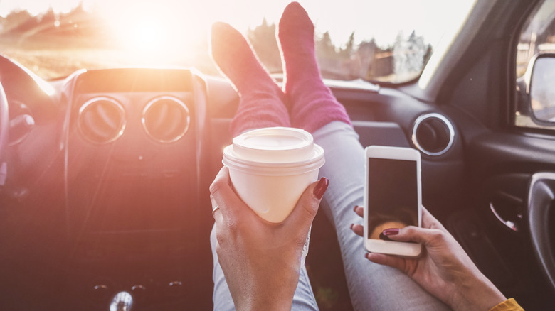 Person with long red nails wearing jeans and purple socks and holding a paper coffee cup and an iphone has their feet propped up on the dashboard of a car. The sun is shining directly into the windshield. 