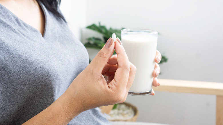 Person holding white pill in one hand and glass of milk in the other