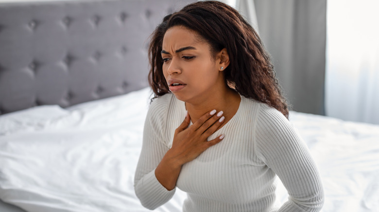 Woman sitting on bed in pain touching a sore throat