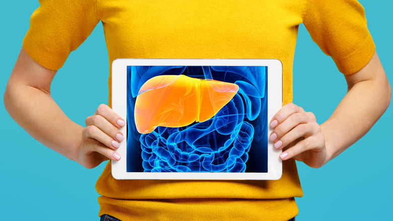 A woman in a yellow shirt holding up a tablet displaying an image of her liver in front of her torso