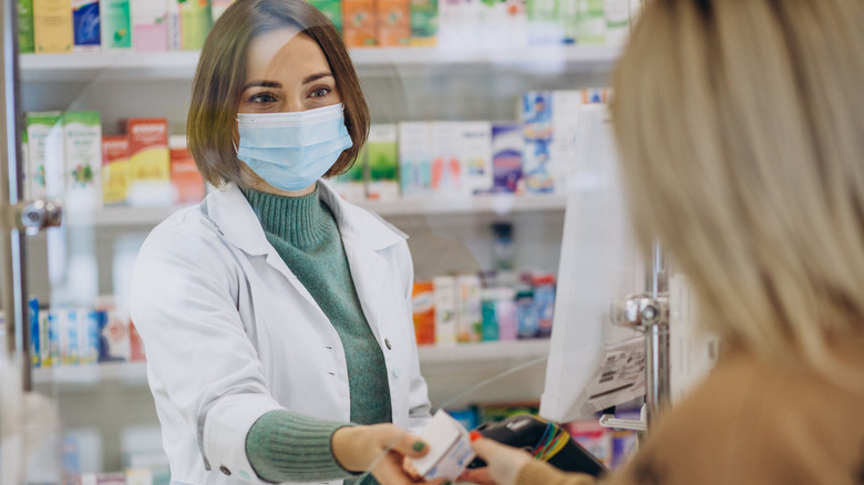 Pharmacist in white coat and face mask handing medication to customer